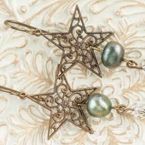 May include: A pair of bronze-colored star-shaped earrings with intricate filigree detailing. Each earring features a green-grey pearl and a clear crystal bead. The earrings are displayed on a textured, off-white surface.