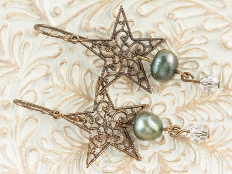 May include: A pair of bronze-colored star-shaped earrings with intricate filigree detailing. Each earring features a green-grey pearl and a clear crystal bead. The earrings are displayed on a textured, off-white surface.