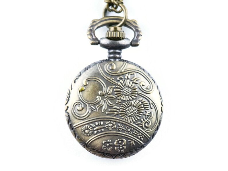 May include: A gold-toned pocket watch with an ornate floral design on the face. The watch has a silver-toned chain and a decorative loop.