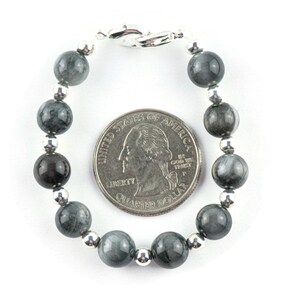 May include: A gray and silver beaded bracelet with a silver clasp. The bracelet is about the size of a US quarter.