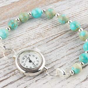 K&ouml;nnte beinhalten: Ein t&uuml;rkisfarbenes und silbernes Perlenarmband mit einer kleinen silbernen Uhr. Das Zifferblatt der Uhr ist wei&szlig; mit schwarzen Zahlen und Zeigern.