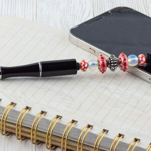 Puede incluir: Un bol&iacute;grafo negro con un clip plateado y un charm de cuentas rojas y blancas. El bol&iacute;grafo est&aacute; sobre un cuaderno con una encuadernaci&oacute;n en espiral dorada. Un tel&eacute;fono inteligente negro est&aacute; en el fondo.