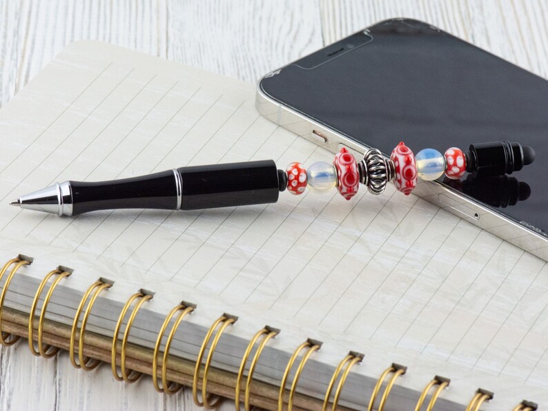 Puede incluir: Un bol&iacute;grafo negro con un clip plateado y un charm de cuentas rojas y blancas. El bol&iacute;grafo est&aacute; sobre un cuaderno con una encuadernaci&oacute;n en espiral dorada. Un tel&eacute;fono inteligente negro est&aacute; en el fondo.