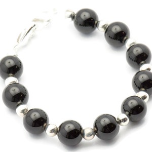 Puede incluir: Un brazalete con grandes cuentas negras brillantes intercaladas con cuentas m&aacute;s peque&ntilde;as de color plateado. El brazalete est&aacute; ensartado con un cierre plateado, creando un dise&ntilde;o simple pero elegante. Las cuentas son redondas y est&aacute;n espaciadas uniformemente.