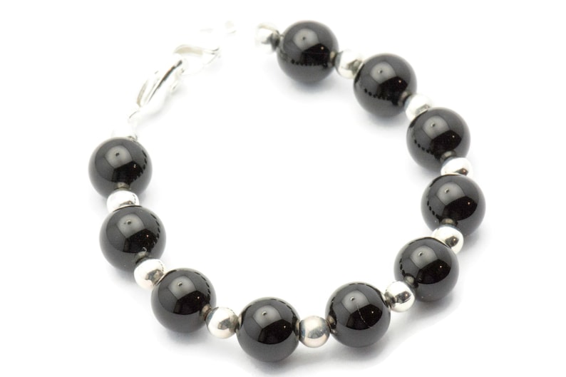 Puede incluir: Un brazalete con grandes cuentas negras brillantes intercaladas con cuentas m&aacute;s peque&ntilde;as de color plateado. El brazalete est&aacute; ensartado con un cierre plateado, creando un dise&ntilde;o simple pero elegante. Las cuentas son redondas y est&aacute;n espaciadas uniformemente.