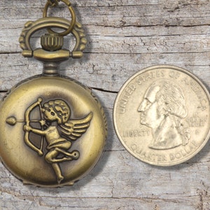 Puede incluir: Un reloj de bolsillo dorado con una cadena y un querub&iacute;n con un arco y alas en la esfera. El reloj est&aacute; al lado de una moneda de 25 centavos de d&oacute;lar estadounidense.
