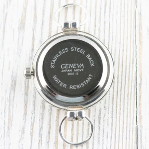 Puede incluir: La parte posterior de un reloj de bolsillo plateado con una esfera negra. La esfera del reloj dice "Geneva Japan Movt 2037-S" y "Stainless Steel Back Water Resistant".