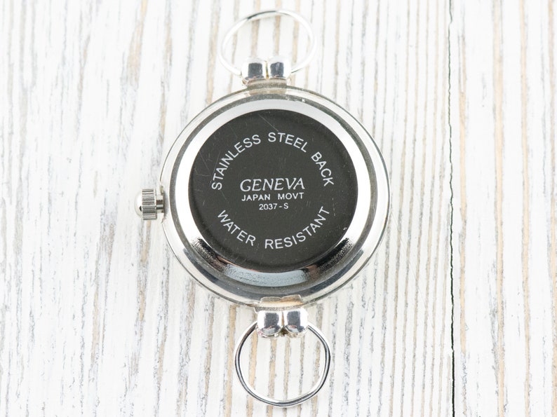 Puede incluir: La parte posterior de un reloj de bolsillo plateado con una esfera negra. La esfera del reloj dice "Geneva Japan Movt 2037-S" y "Stainless Steel Back Water Resistant".