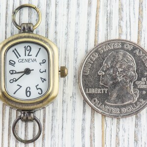 Pu&ograve; includere: Un piccolo orologio da tasca in stile antico con un quadrante quadrato e numeri romani. L'orologio ha una finitura in ottone e un quadrante bianco con lancette nere. L'orologio &egrave; appeso a una catena argentata.