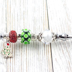 May include: A silver bottle opener with Christmas-themed beads. The beads include a red, sparkly bead, a green and white polka-dot bead, and a clear, faceted bead. A small Christmas tree charm hangs from the opener.