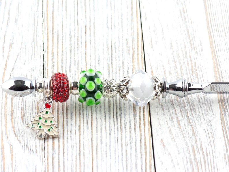 May include: A silver bottle opener with Christmas-themed beads. The beads include a red, sparkly bead, a green and white polka-dot bead, and a clear, faceted bead. A small Christmas tree charm hangs from the opener.