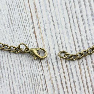 K&ouml;nnte beinhalten: Nahaufnahme einer bronzefarbenen Halskette mit Karabinerverschluss. Die Kette besteht aus kleinen, miteinander verbundenen Gliedern. Der Verschluss ist ge&ouml;ffnet und zeigt den Verbindungspunkt. Der Hintergrund ist eine helle, verwitterte Holzoberfl&auml;che.