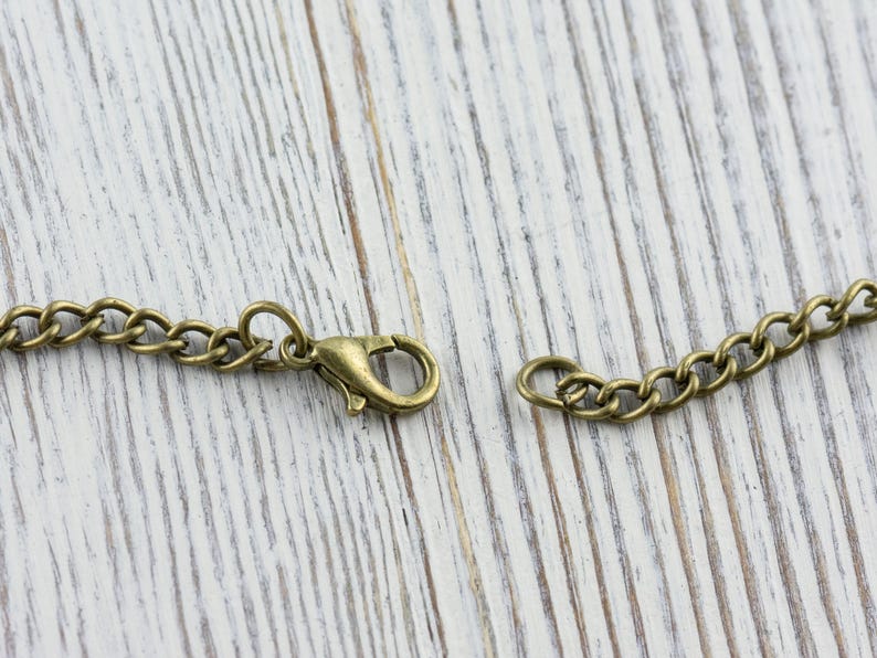 K&ouml;nnte beinhalten: Nahaufnahme einer bronzefarbenen Halskette mit Karabinerverschluss. Die Kette besteht aus kleinen, miteinander verbundenen Gliedern. Der Verschluss ist ge&ouml;ffnet und zeigt den Verbindungspunkt. Der Hintergrund ist eine helle, verwitterte Holzoberfl&auml;che.