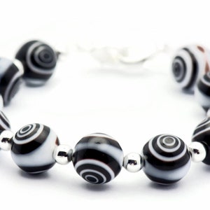 K&ouml;nnte beinhalten: Ein schwarz-wei&szlig;es Armband mit runden, gemusterten Perlen. Die Perlen haben ein wirbelndes, konzentrisches Kreisdesign.
