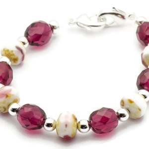 K&ouml;nnte beinhalten: Ein Armband mit abwechselnd wei&szlig;en und burgunderfarbenen Perlen. Die wei&szlig;en Perlen haben ein Blumenmuster. Das Armband hat einen silbernen Verschluss.
