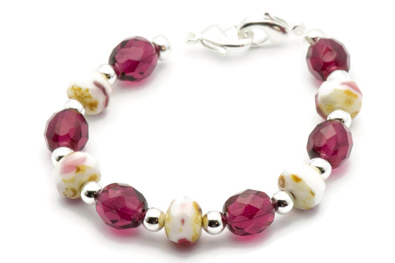 K&ouml;nnte beinhalten: Ein Armband mit abwechselnd wei&szlig;en und burgunderfarbenen Perlen. Die wei&szlig;en Perlen haben ein Blumenmuster. Das Armband hat einen silbernen Verschluss.
