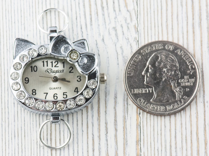 May include: A silver-tone watch with a white face and rhinestone accents. The watch is shaped like a cat's face with a bow on top. The watch is next to a US quarter for size comparison.