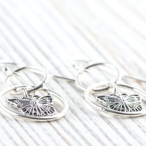 May include: Silver butterfly earrings. Each earring features a detailed butterfly charm suspended within a circular frame. The earrings are designed with a hook closure and are displayed on a white wooden surface.