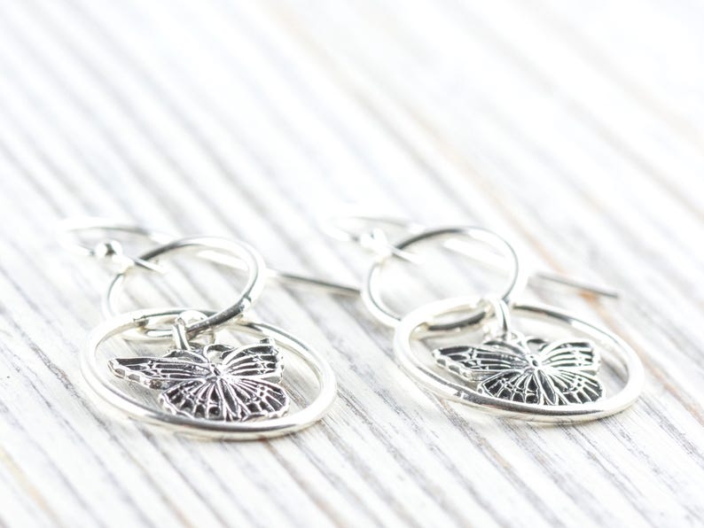 May include: Silver butterfly earrings. Each earring features a detailed butterfly charm suspended within a circular frame. The earrings are designed with a hook closure and are displayed on a white wooden surface.