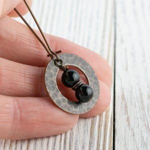Peut inclure: Boucle d'oreille artisanale avec un pendentif ovale en m&eacute;tal martel&eacute; et deux perles noires. La boucle d'oreille a un fil de couleur bronze et est tenue dans une main sur un fond en bois blanc. Le bijou est simple et &eacute;l&eacute;gant.