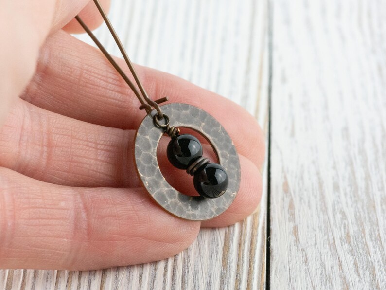 Peut inclure: Boucle d'oreille artisanale avec un pendentif ovale en m&eacute;tal martel&eacute; et deux perles noires. La boucle d'oreille a un fil de couleur bronze et est tenue dans une main sur un fond en bois blanc. Le bijou est simple et &eacute;l&eacute;gant.