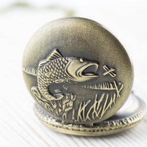 Pode incluir: Um rel&oacute;gio de bolso de bronze com uma grava&ccedil;&atilde;o detalhada de um peixe saltando para fora da &aacute;gua. O mostrador do rel&oacute;gio n&atilde;o &eacute; vis&iacute;vel.