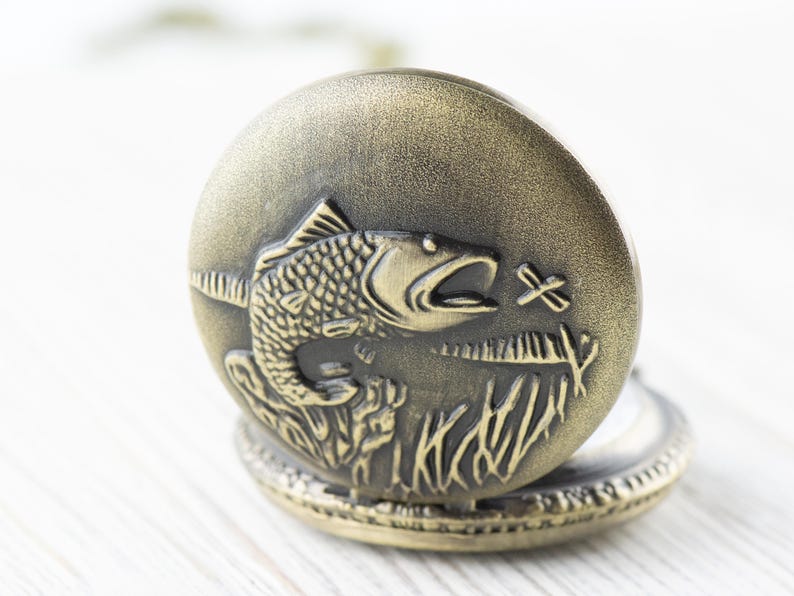 Pode incluir: Um rel&oacute;gio de bolso de bronze com uma grava&ccedil;&atilde;o detalhada de um peixe saltando para fora da &aacute;gua. O mostrador do rel&oacute;gio n&atilde;o &eacute; vis&iacute;vel.