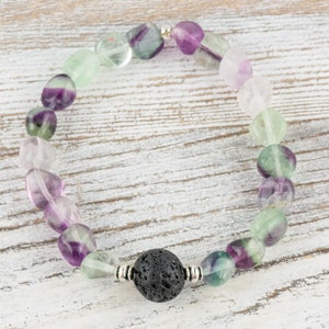 Puede incluir: Un brazalete hecho de cuentas de fluorita verde, p&uacute;rpura y blanca con una cuenta de lava negra en el centro.