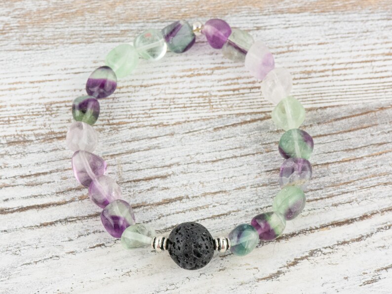 Puede incluir: Un brazalete hecho de cuentas de fluorita verde, p&uacute;rpura y blanca con una cuenta de lava negra en el centro.