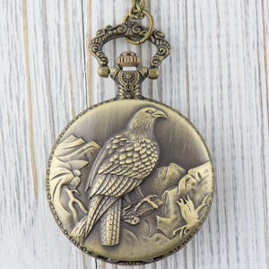 May include: A bronze pocket watch with a detailed engraving of a bird on the front. The watch has a chain attached to the top.