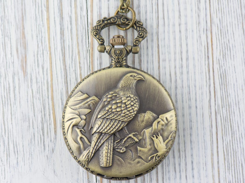 May include: A bronze pocket watch with a detailed engraving of a bird on the front. The watch has a chain attached to the top.