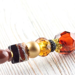 Puede incluir: Primer plano de un objeto decorativo con cuentas. Incluye un extremo puntiagudo marr&oacute;n, un cuadrado con patrones marrones y negros, una esfera dorada y gemas facetadas de color &aacute;mbar y naranja rojizo. Tambi&eacute;n hay un acento floral dorado.