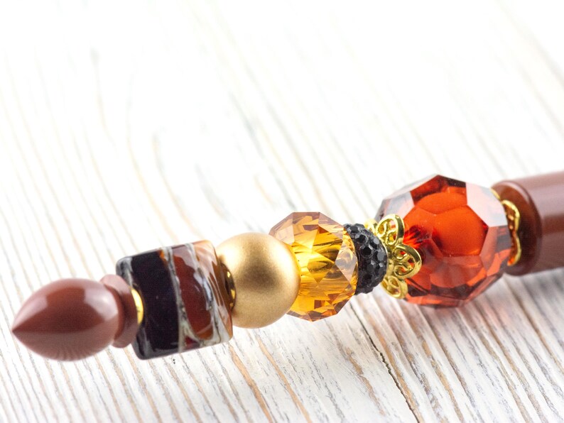 Puede incluir: Primer plano de un objeto decorativo con cuentas. Incluye un extremo puntiagudo marr&oacute;n, un cuadrado con patrones marrones y negros, una esfera dorada y gemas facetadas de color &aacute;mbar y naranja rojizo. Tambi&eacute;n hay un acento floral dorado.