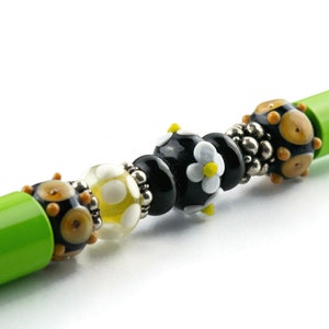 May include: A green pen with a black, white, and yellow glass bead design. The beads are in a pattern of black, white, yellow, black, and then a black bead with a white flower with a yellow center.