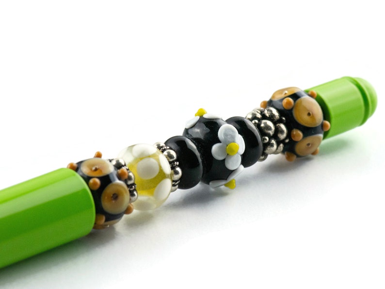 May include: A green pen with a black, white, and yellow glass bead design. The beads are in a pattern of black, white, yellow, black, and then a black bead with a white flower with a yellow center.