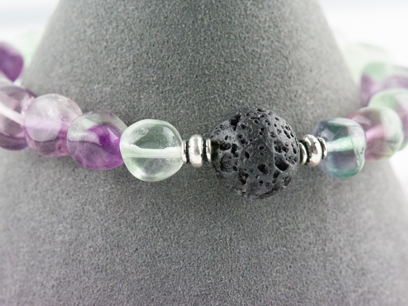 Puede incluir: Un brazalete hecho con cuentas de fluorita verde, p&uacute;rpura y blanca y una cuenta de roca de lava negra. El brazalete tiene cuentas espaciadoras plateadas.