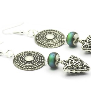May include: Silver earrings with a round, ornate charm, a green iridescent bead, and a triangular charm with a filigree design.