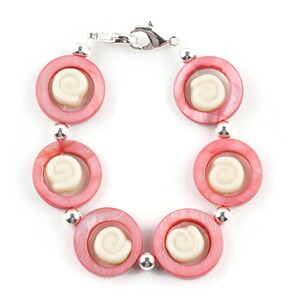 Pu&ograve; includere: Un braccialetto rosa e bianco con una chiusura argentata. Il braccialetto &egrave; fatto di perline rotonde con un design a spirale bianco al centro di ogni perla.