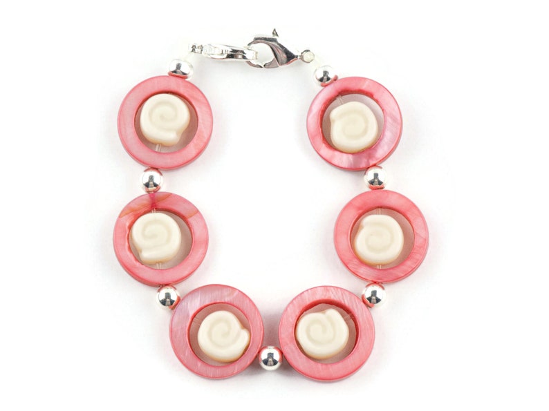 Pu&ograve; includere: Un braccialetto rosa e bianco con una chiusura argentata. Il braccialetto &egrave; fatto di perline rotonde con un design a spirale bianco al centro di ogni perla.