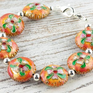 May include: A bracelet with a silver clasp and orange, green, and white enamel beads with floral designs.