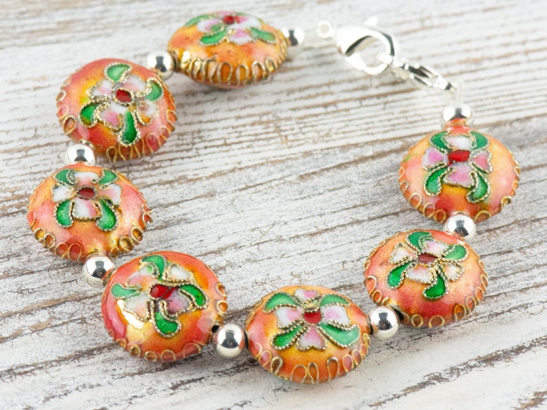 May include: A bracelet with a silver clasp and orange, green, and white enamel beads with floral designs.