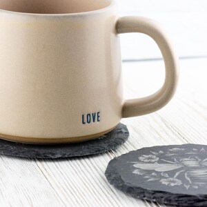 May include: A beige ceramic mug with the word "LOVE" printed on the side, resting on a dark gray slate coaster. Another slate coaster with a bee and floral design is next to it. The mug has a large handle.