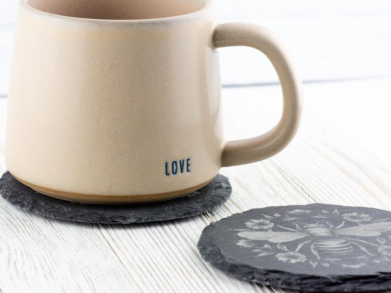 May include: A beige ceramic mug with the word "LOVE" printed on the side, resting on a dark gray slate coaster. Another slate coaster with a bee and floral design is next to it. The mug has a large handle.