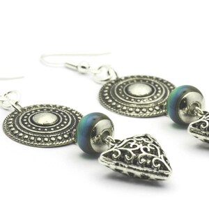 May include: Silver earrings with a round, patterned disc, a blue and green iridescent bead, and a triangular, filigree charm.