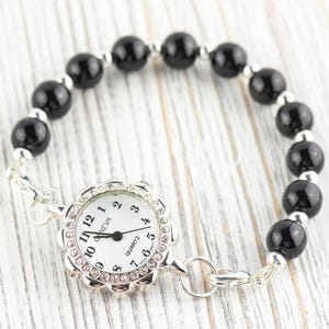 May include: A silver-toned bracelet watch with a white face and black hands. The watch face is surrounded by a floral-shaped bezel with clear crystals. The band features black, round beads and silver-toned spacers.