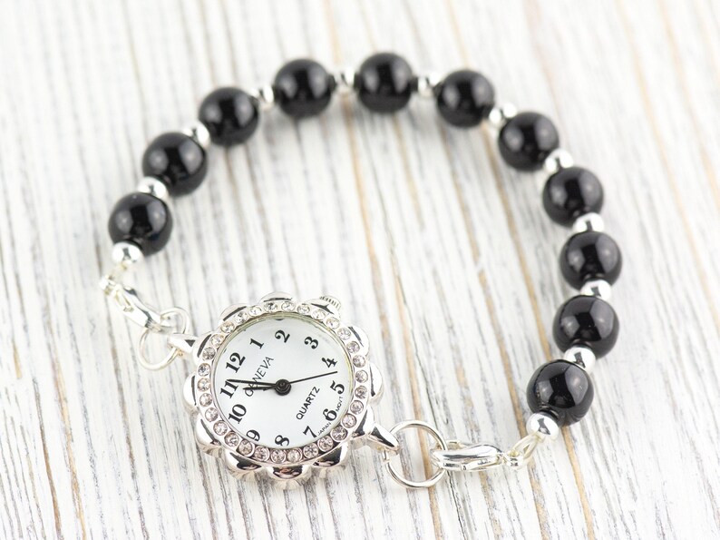 May include: A silver-toned bracelet watch with a white face and black hands. The watch face is surrounded by a floral-shaped bezel with clear crystals. The band features black, round beads and silver-toned spacers.