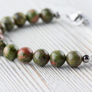 Puede incluir: Un brazalete de piedras preciosas verdes y rojas con un cierre plateado. El brazalete est&aacute; hecho de cuentas redondas y est&aacute; sobre una superficie de madera blanca.