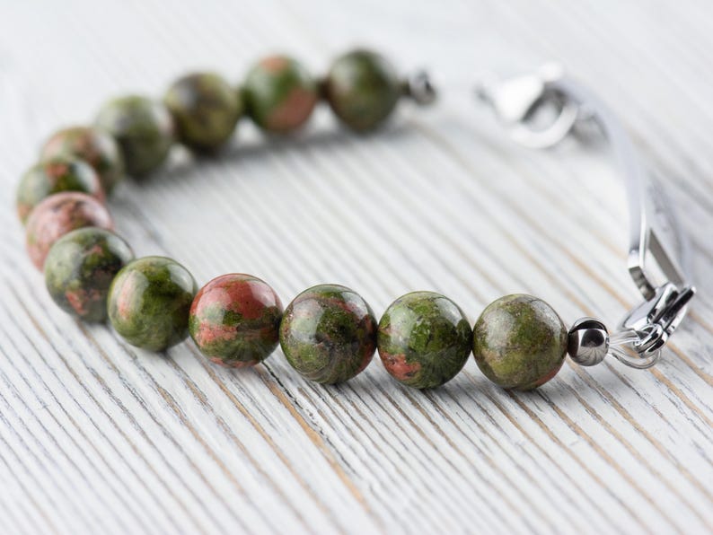 Puede incluir: Un brazalete de piedras preciosas verdes y rojas con un cierre plateado. El brazalete est&aacute; hecho de cuentas redondas y est&aacute; sobre una superficie de madera blanca.