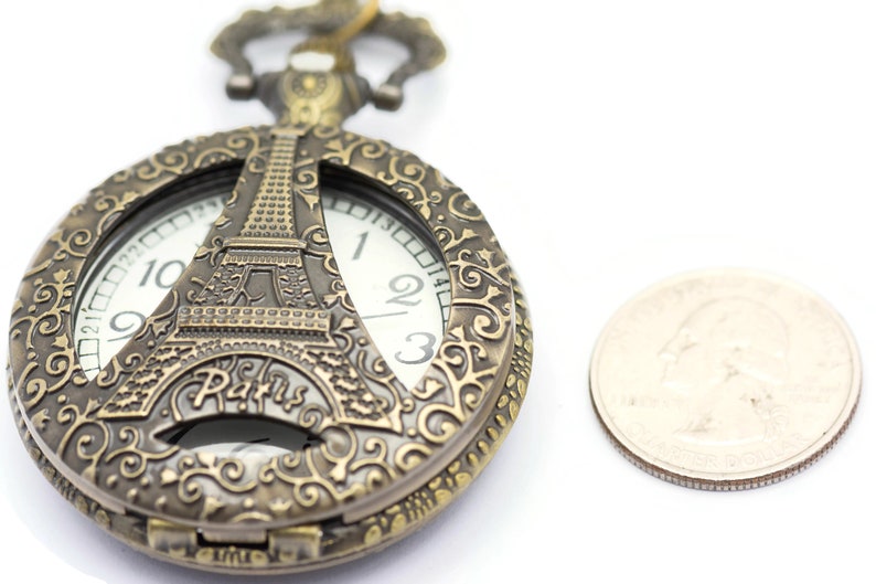 Może przedstawiać: Złoty zegarek kieszonkowy z ozdobnym wzorem przedstawiającym wieżę Eiffla i napis "Paris". Tarcza zegarka jest widoczna z rzymskimi cyframi.