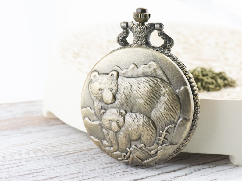 May include: A bronze pocket watch with a detailed design of a mother bear and her cub. The watch has a decorative chain attached.